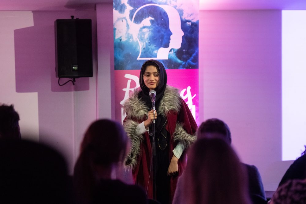 A young woman stands in front of a microphone, a row of people sit watching her on stage.