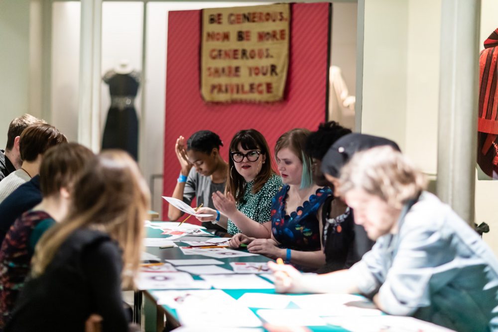 Led by a workshop leader, young people sit around a table in a room with artworks.