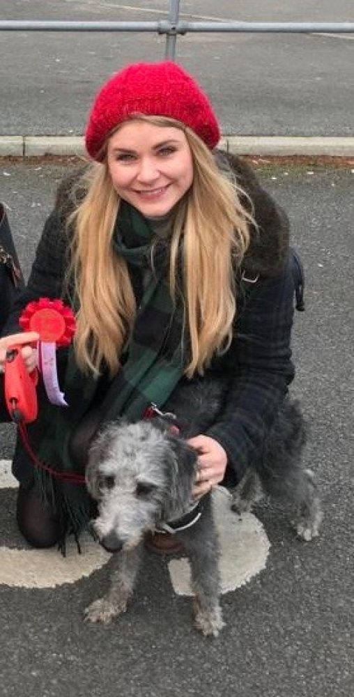 A blonde woman in a red hat crouches on the street holding a red rosette and a grey dog.