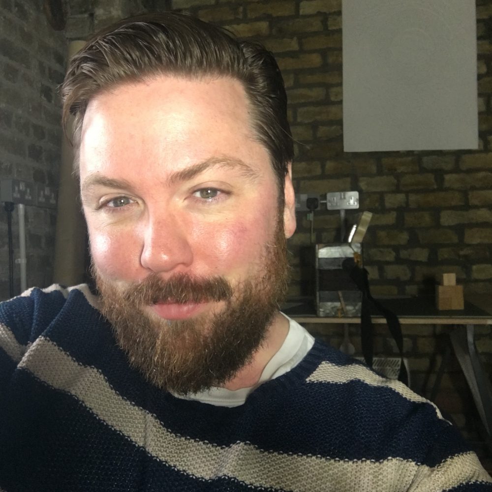 A head and shoulders view of a man wearing a stripey top with a desk and brick wall behind him.