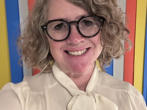 A woman with shoulder length wavy blonde hair and black rimmed spectacles smiling wearing a cream blouse