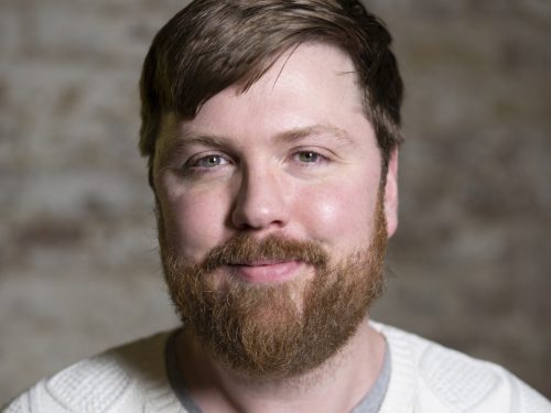 A head and shoulders view of a man with a beard wearing a white jumper