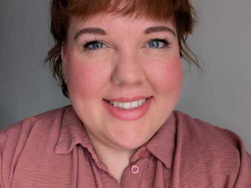 A photo of a female with tied back auburn hair and a fringe. They are smiling and wearing a dusky pink shirt.