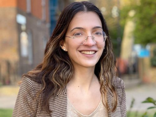 A person with long brown hair and glasses wearing a beige top and blazer, smiling and standing outdoors