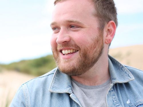 A man wearing a denim jacket smiles on a beach