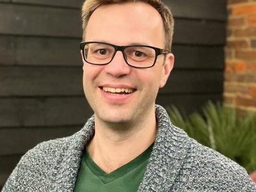 A man stands in front of a wood pannelled wall. He wears glasses, a grey cardigan and green tshirt