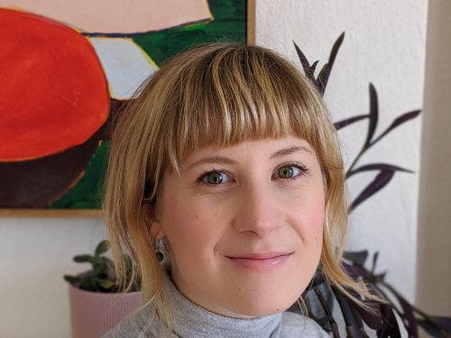A head and shoulders view of a woman wearing a grey turtleneck, standing in front of a painting and two house plants.