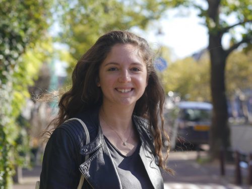 A woman in a leather jacket stands in the centre of a path on a tree lined street.