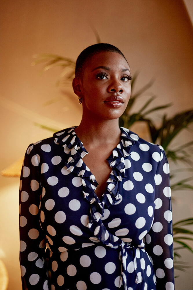 A woman in a black and white polka dot dress stands outside with plants behind her.