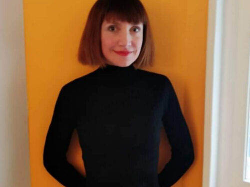 A woman wearing a black turtleneck stands in front of an orange wall