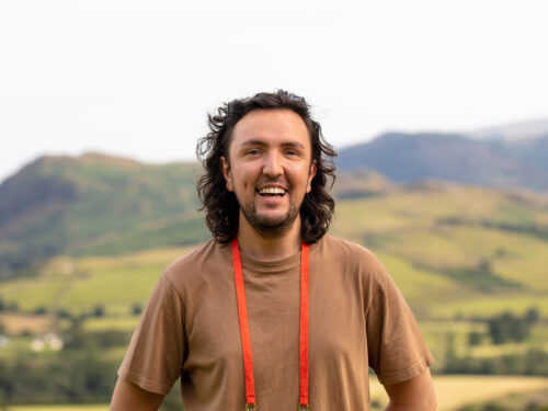 A man with shoulder length brown hair and wearing a brown t-shirt. They are smiling, in the background is rolling hills