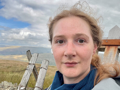 Head and shoulders of a woman with tied back hair wearing an anorak. In the background is hills