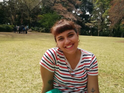A woman sitting on grass on a sunny day. She is smiling and wearing a striped t-shirt and jeans