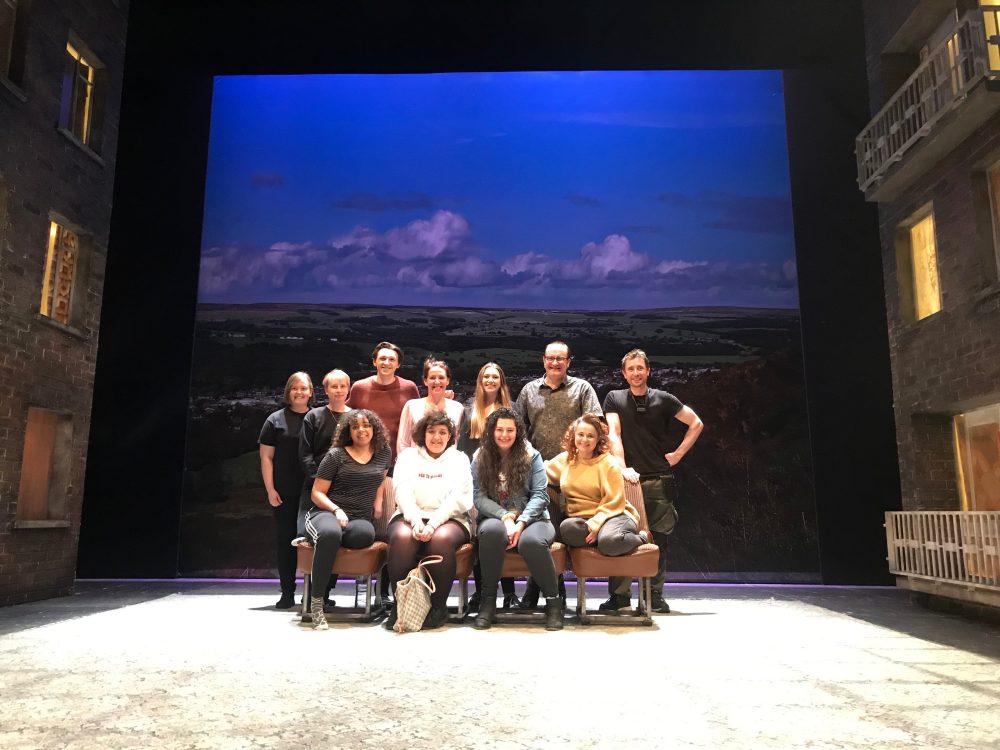 A group of 11 people sit and stand on the stage of a theatre, with a backdrop of blue clouds behind them