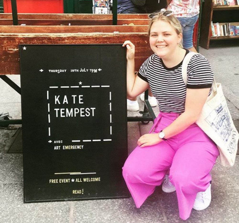A young woman crouches next to a black sign which reads Kate Tempest avec Arts Emergency. Free event! All welcome! Read!