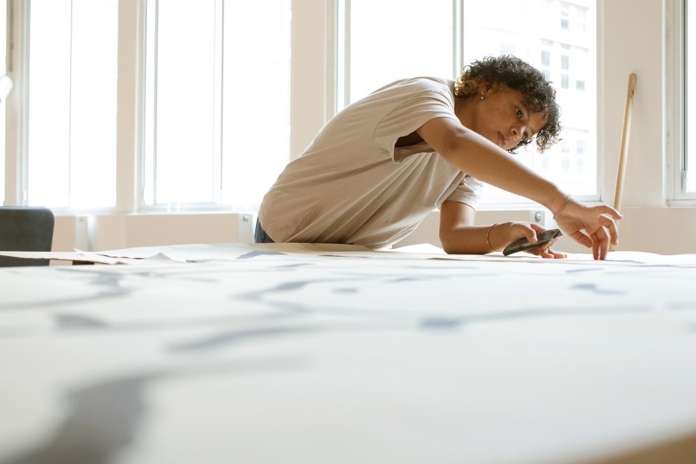 A black girl in a beige t-shirt with short hair draws on a large flat piece of set for a play.