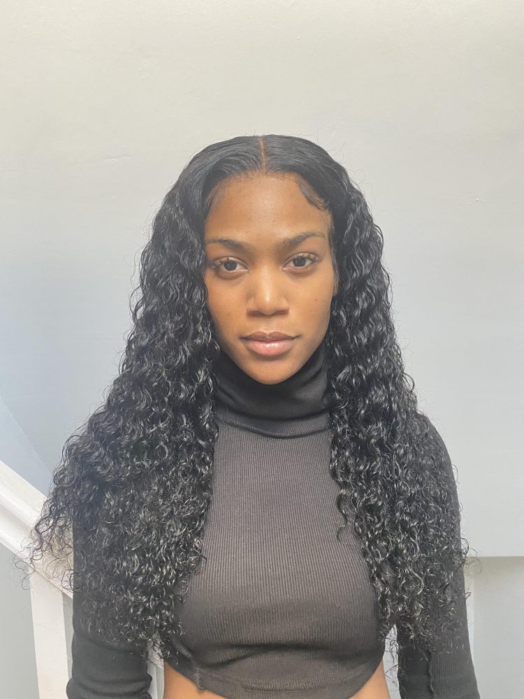 A young woman looks straight at the camera, she wears a brown polo neck and stands in front of a white wall