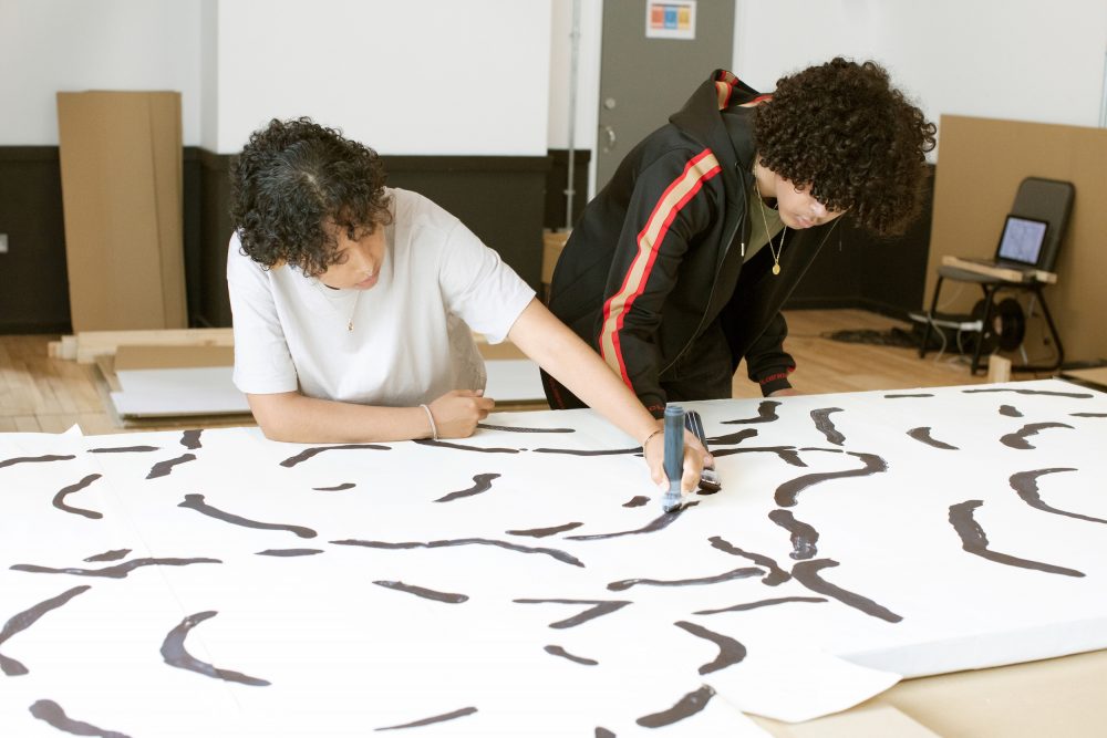 A black young woman in a beige t-shirt and a black young man wearing a black sweatshirt draw on a large flat piece of set for a play.