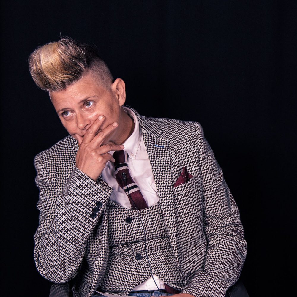 A woman in a herringbone suit sits and has her hand over her mouth