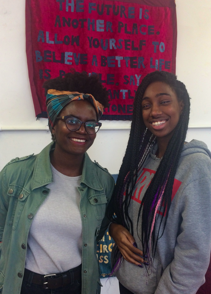 Two women stand under an Arts Emergency banner that reads 'The future is another place. Allow yourself to believe a better world is possible...'. On the left is Renni Eddo-Lodge. She is the shorter of the two, has dark brown skin, curly dark hair tied up with a bright scarf, and is wearing a white t-shirt, khaki overshirt, and glasses. On the right is Sile Edwards. She also has dark brown skin and long braided dark hair in down to her waist. Her hair has a pink braid in it, creating a streak. She is wearing a grey hoodie with the Marvel Comics logo on it in bright red. They are both smiling at the camera.