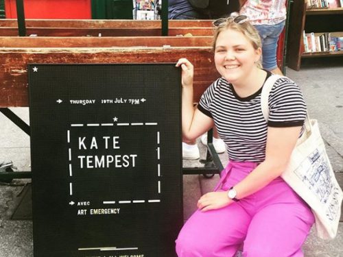 A young woman crouches next to a black sign which reads Kate Tempest avec Arts Emergency. Free event! All welcome! Read!