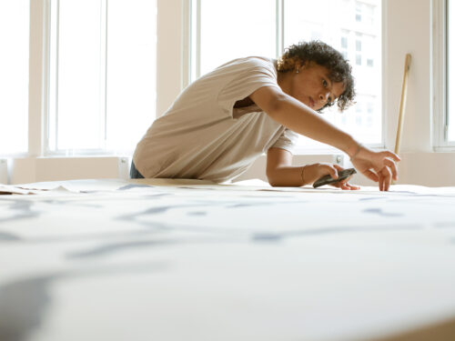 A black girl in a beige t-shirt with short hair draws on a large flat piece of set for a play.