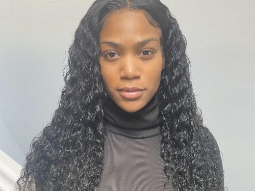 A young woman looks straight at the camera, she wears a brown polo neck and stands in front of a white wall