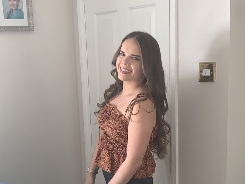 A young woman with long brown hair stands in front of a door.