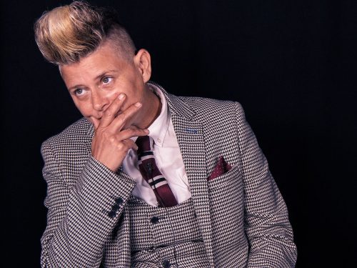 A woman in a herringbone suit sits and has her hand over her mouth