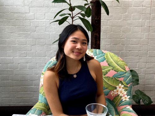A young woman sits at a table with a plant and brick wall behind her.