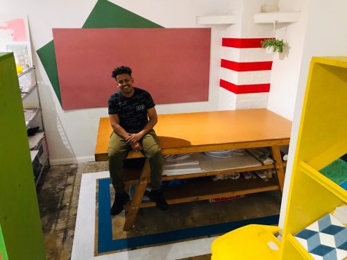 A young man sits on a table in a room full of colour shapes and furniture