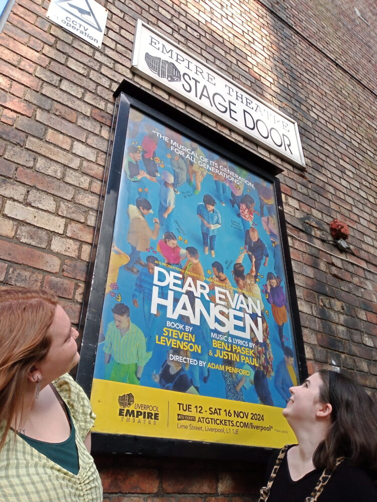 2 women, 1 auburn haired wearing a green top, 1 dark haired wearing a black top. They are outside and looking at a theatre poster for the show 'Dear Evan Hansen'.
