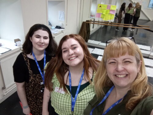 Three women; 1 dark haired wearing a black top, 1 auburn haired wearing a green checked top and 1 blonde haired wearing a black top, smiling to camera. They are Youth Collective member Amie, Young Community member Beth and Arts Emergency Head of Community Rose, at Liverpool Empire Theatre